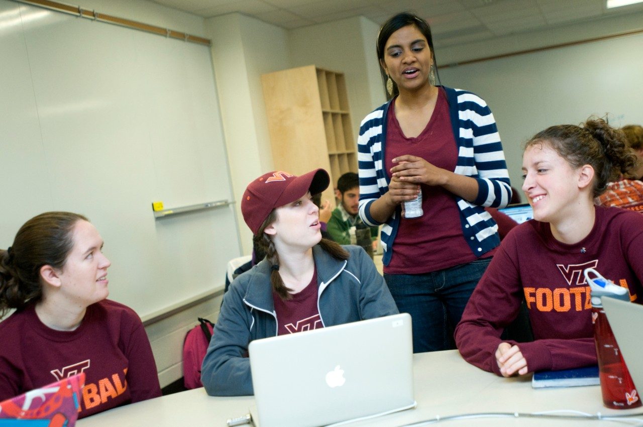 3 students sitting, 1 standing around a table with a laptop